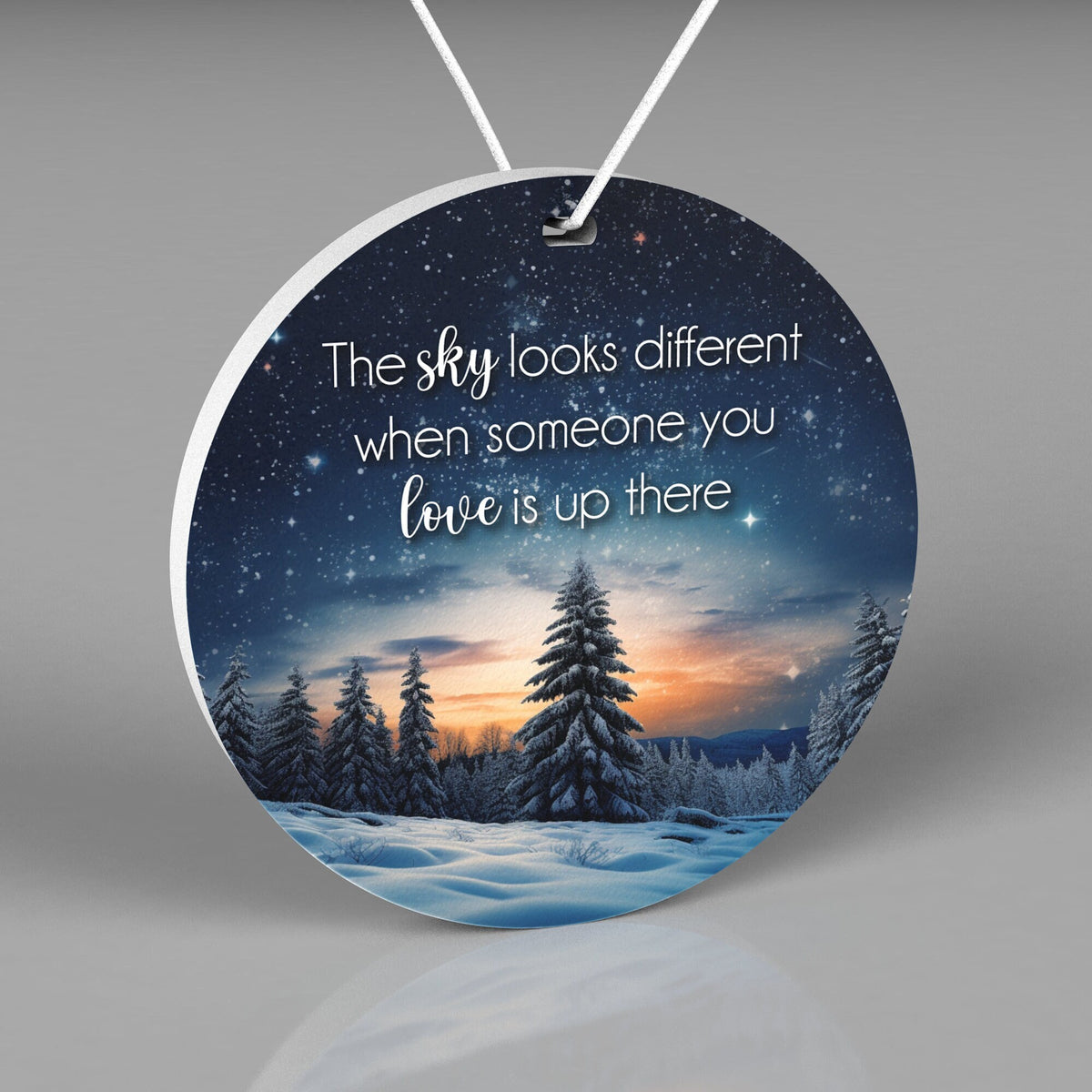 Round Christmas ornament with a night sky, stars, snow and pine trees Words say "The sky looks different when someone you love is up there"